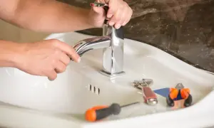 Ręce majsterkowicza naprawiają cieknący kran dwuuchwytowy. Obok leżą klucz, śrubokręt i kombinerki.
