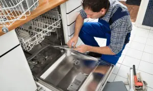 Instalator w niebieskim kombinezonie pokazuje, jak podpiąć zmywarkę, pracując przy jej wnętrzu. Narzędzia leżą obok.