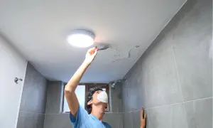 Mężczyzna w masce usuwa łopatką łuszczącą się farbę z sufitu w łazience.