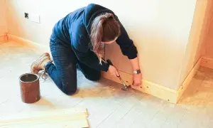 Kobieta przybija młotkiem listwy przypodłogowe. Widać puszkę i deski, co sugeruje, czym przykleić listwy przypodłogowe.