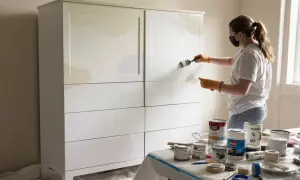 Kobieta w masce i rękawiczkach maluje białą farbą szafę. Na stole puszki z farbą i akcesoria, jak pomalować meble na biało.
