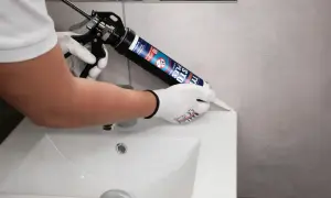 Ręce w rękawiczkach nakładają biały silikon sanitarny z pistoletu na krawędź umywalki, uszczelniając połączenie.