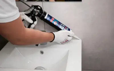 Ręce w rękawiczkach nakładają czarny silikon sanitarny na krawędź umywalki, uszczelniając połączenie.