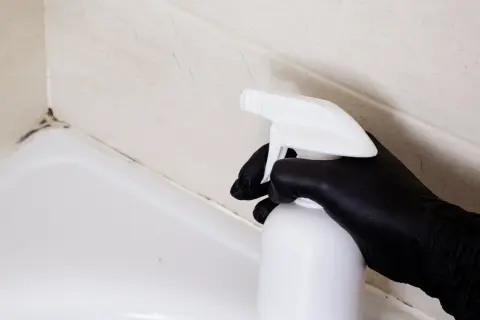 Czarna rękawiczka trzyma biały spray, gotowy do działania. Czas na czyszczenie silikonu w kabinie prysznicowej i pozbycie się pleśni.