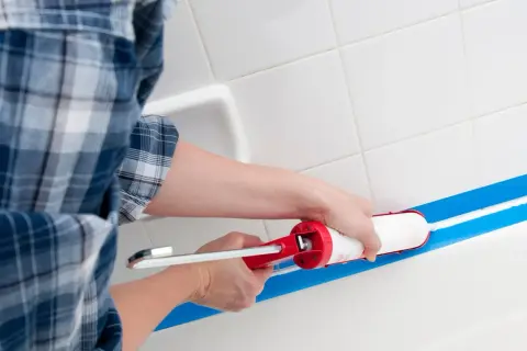 Osoba w kratę zakłada silikon sanitarny w wannie, używając pistoletu i taśmy malarskiej.