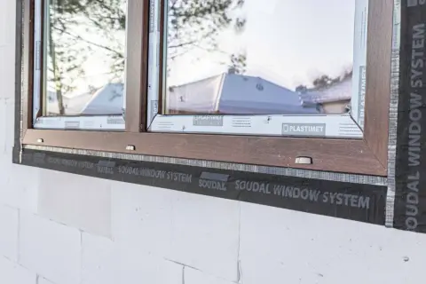 Brązowe okno w trakcie ciepłego montażu z użyciem taśmy SOUDAL WINDOW SYSTEM. Widoczna biała ściana z bloczków.