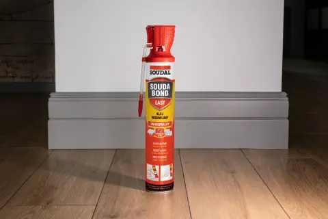 Puszka kleju budowlanego Soudal Easy Bond na drewnianej podłodze, obok szarej listwy przypodłogowej.