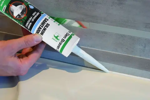 Ręka trzyma tubkę silikonu sanitarnego, aplikując go wzdłuż krawędzi zlewu, pokazując jak uszczelnić zlew.