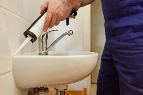Ręce nakładają silikon wokół umywalki w łazience. Dowiedz się, jak wymienić silikony w łazience, aby zapewnić szczelność.