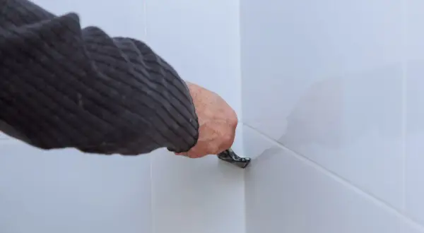 Ręka w swetrze usuwa silikon z białych paneli podłogowych za pomocą narzędzia.