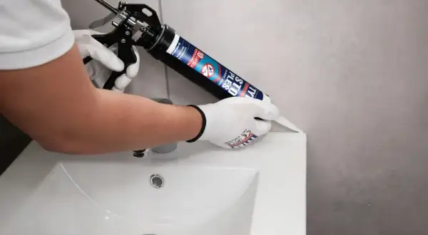 Ręce w rękawiczkach nakładają czarny silikon sanitarny na krawędź umywalki, uszczelniając połączenie.