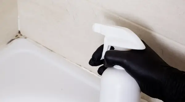 Czarna rękawiczka trzyma biały spray, gotowy do działania. Czas na czyszczenie silikonu w kabinie prysznicowej i pozbycie się pleśni.