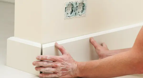 Ręce z pyłem gipsowym przyklejają białe listwy podłogowe do ściany. W tle widać gniazdka elektryczne.