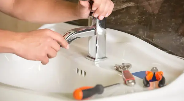 Ręce majsterkowicza naprawiają kapiący kran. Obok leżą klucz, śrubokręt i kombinerki.