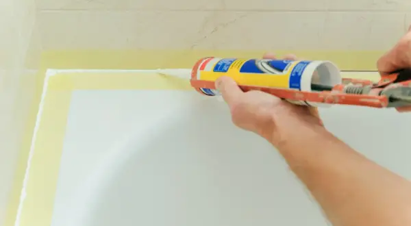 Aplikacja silikonu do brodzika. Ręka trzymająca pistolet z silikonem, nakładająca białą masę na krawędź brodzika.