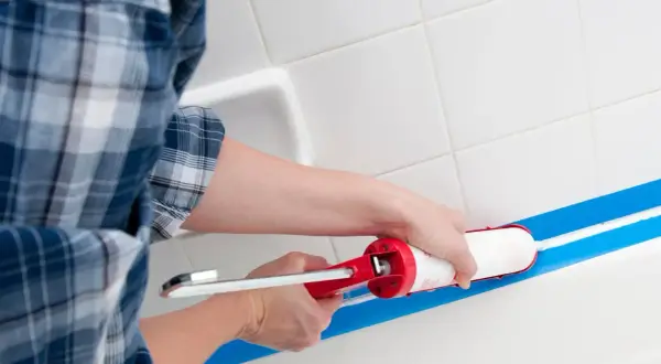 Osoba w kratę zakłada silikon sanitarny w wannie, używając pistoletu i taśmy malarskiej.