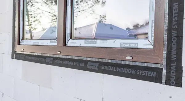 Brązowe okno w trakcie ciepłego montażu z użyciem taśmy SOUDAL WINDOW SYSTEM. Widoczna biała ściana z bloczków.