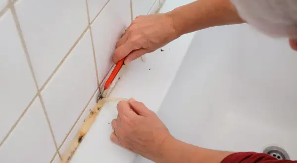 Ręce usuwają stary, zabrudzony silikon z wanny za pomocą nożyka. Dowiedz się, jak usunąć silikon z materiału, aby odnowić łazienkę.