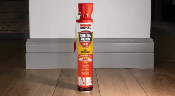 Puszka kleju budowlanego Soudal Easy Bond na drewnianej podłodze, obok szarej listwy przypodłogowej.