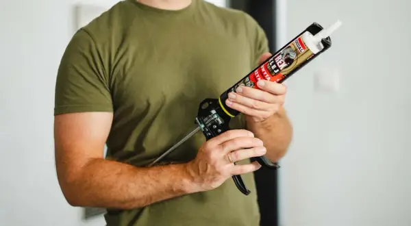 Mężczyzna w zielonej koszulce trzyma pistolet z tubą kleju do listew przypodłogowych plastikowych.