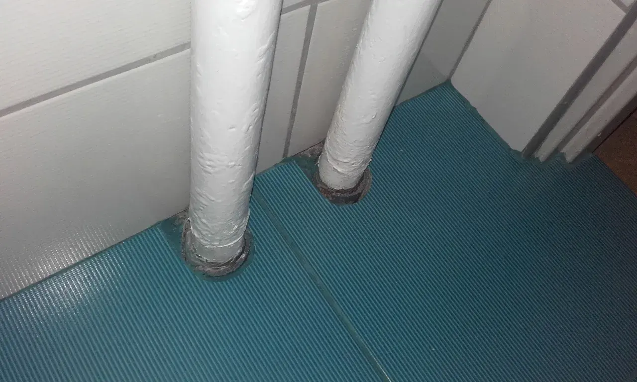 Dwa białe pionowe rury wychodzące z podłogi, przykryte zieloną wykładziną. Pomysł, jak zamaskować rury co przy podłodze.