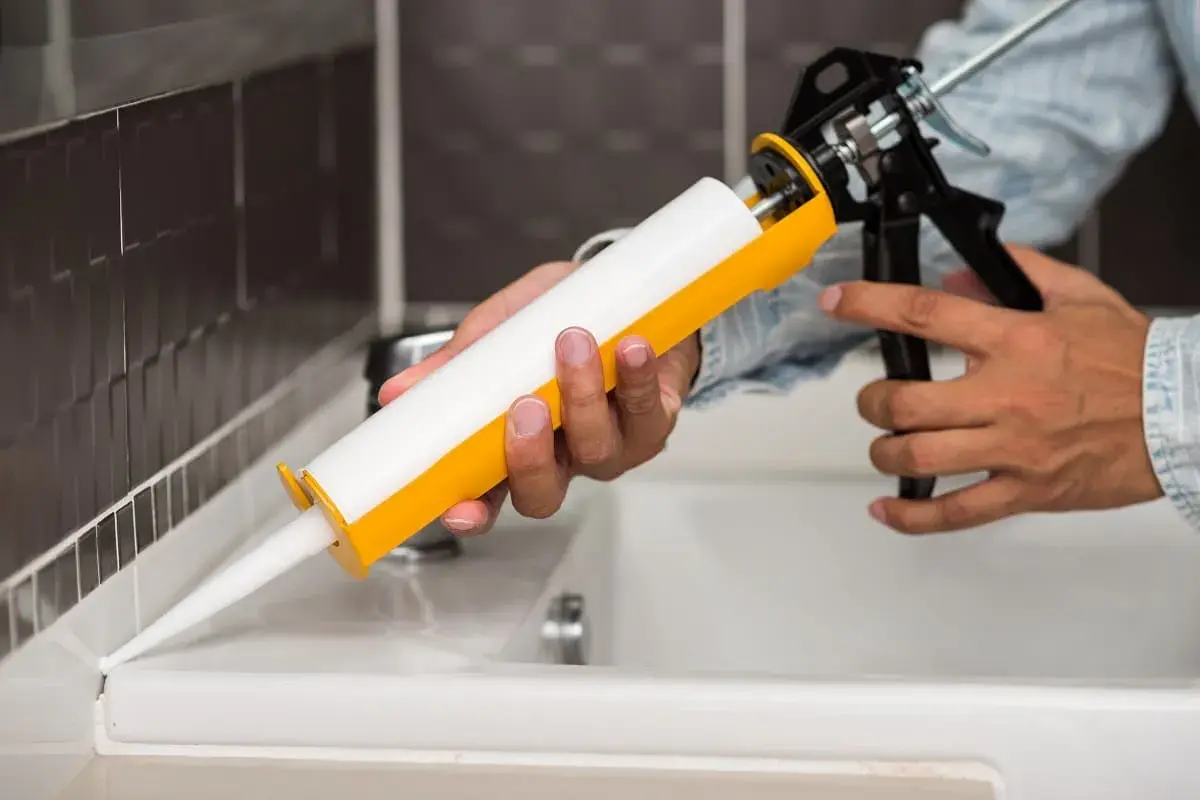 Dłoń trzymająca pistolet do silikonu, aplikująca biały uszczelniacz na biały blat przy zlewie.