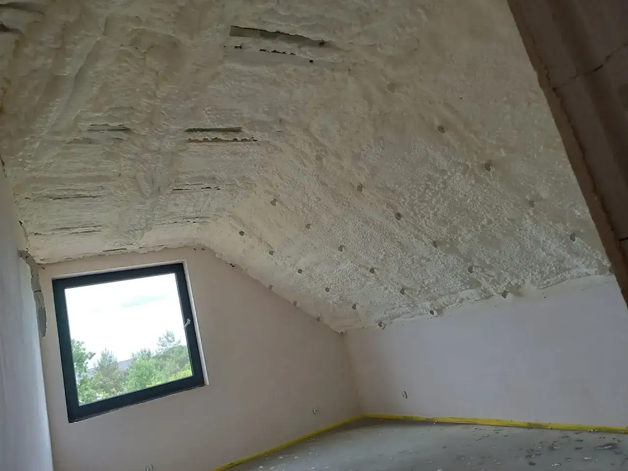 Pianka pur w sprayu izoluje poddasze. Widoczny dach skośny pokryty pianką, okno i ściany.