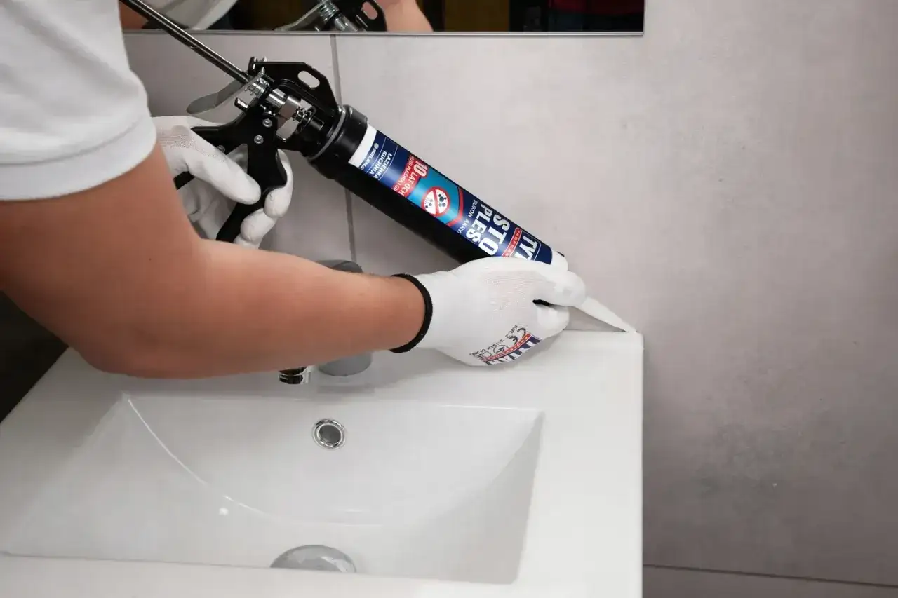 Ręce w rękawiczkach nakładają biały silikon sanitarny z pistoletu na krawędź umywalki, uszczelniając połączenie.