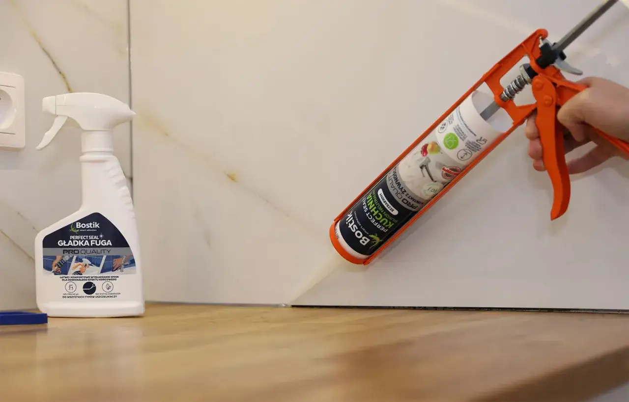 Aplikacja silikonu do blatu kuchennego. Pistolet z tubą silikonu Bostik Perfect Seal Kuchnia neutralny, obok butelka preparatu do wygładzania fug.