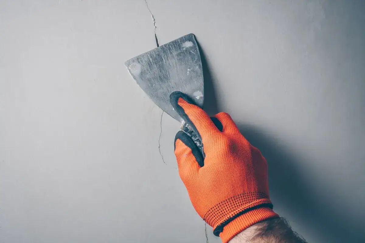 Dłoń w pomarańczowej rękawicy zacierająca pęknięcie na ścianie szpachlą. Użyto akryl lekki szpachlowy.