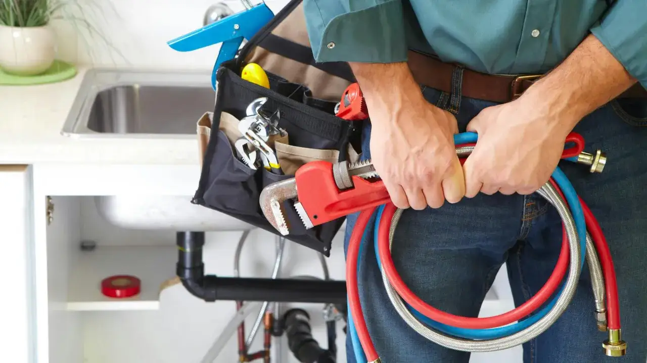 Hydraulik z torbą narzędziową i kluczem nastawnym przygotowuje się do pracy. Pokazuje, jak wykręcić wężyki z baterii kuchennej.