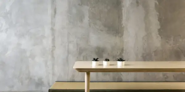 Minimalistyczne wnętrze z surową, betonową ścianą. Na jasnym drewnianym stole stoją trzy małe rośliny w białych doniczkach. Pytanie: czy beton architektoniczny można malować?