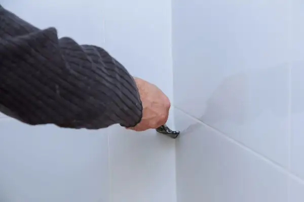 Ręka w swetrze usuwa silikon z białych paneli podłogowych za pomocą narzędzia.
