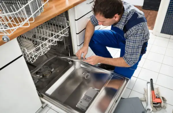 Instalator w niebieskim kombinezonie pokazuje, jak podpiąć zmywarkę, pracując przy jej wnętrzu. Narzędzia leżą obok.