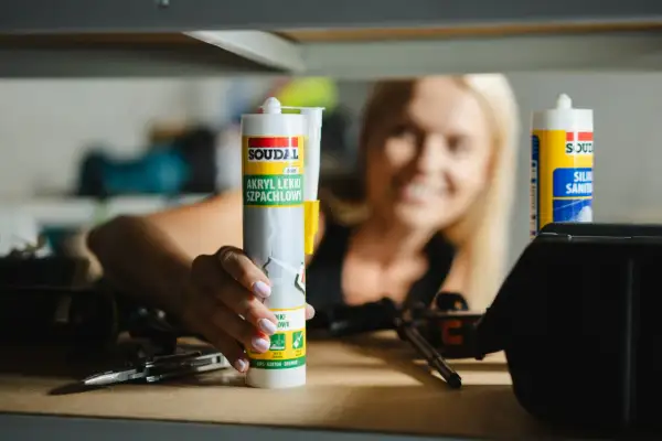 Kobieta trzyma tubę akrylu lekkiego szpachlowego Soudal. Czas schnięcia silikonu akrylowego zależy od warunków.