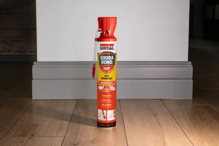 Puszka kleju budowlanego Soudal Easy Bond na drewnianej podłodze, obok szarej listwy przypodłogowej.